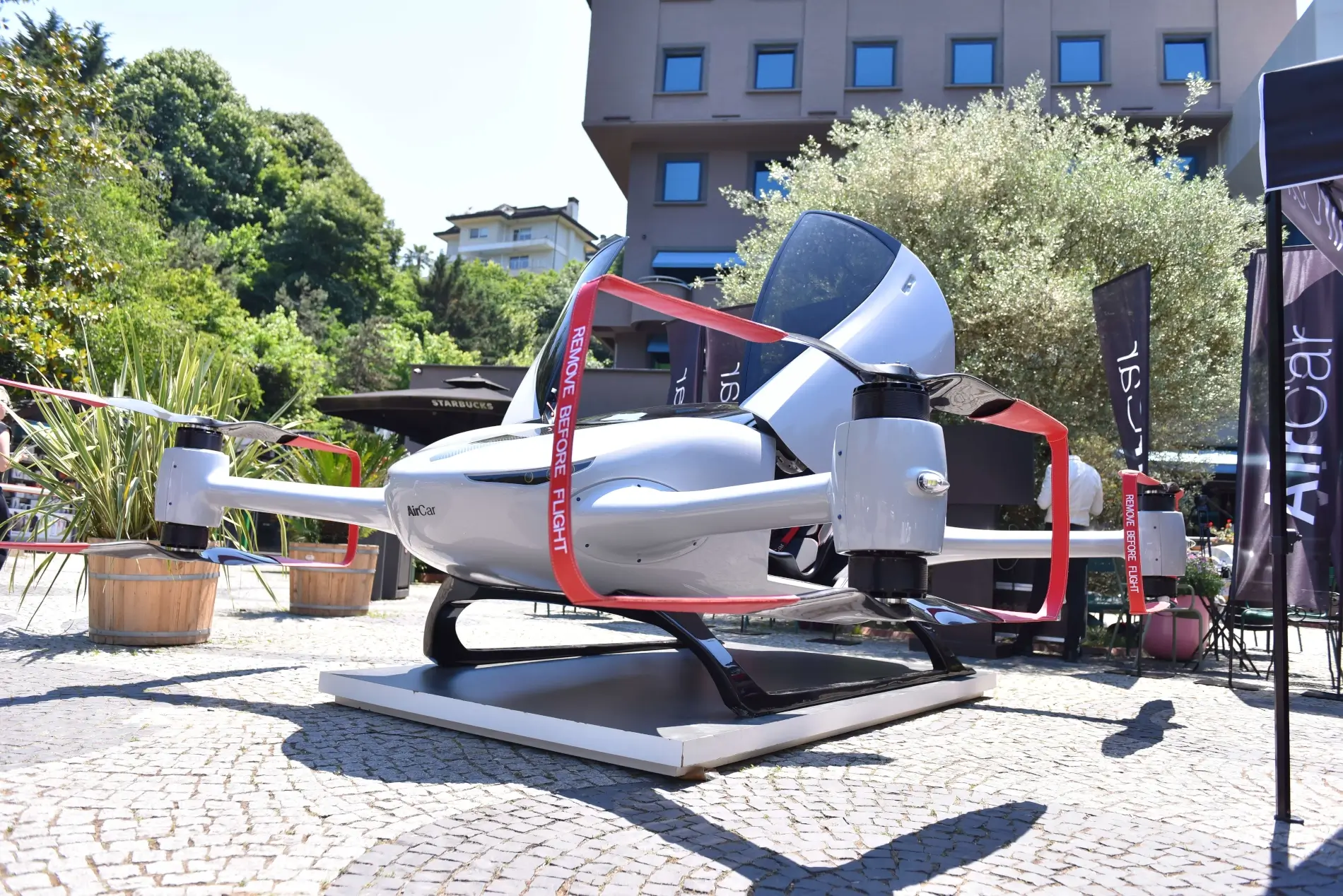 Boční pohled na zaparkovaný bílý letoun AirCar s vyklopenými dveřmi a rotory na městské prezentaci.