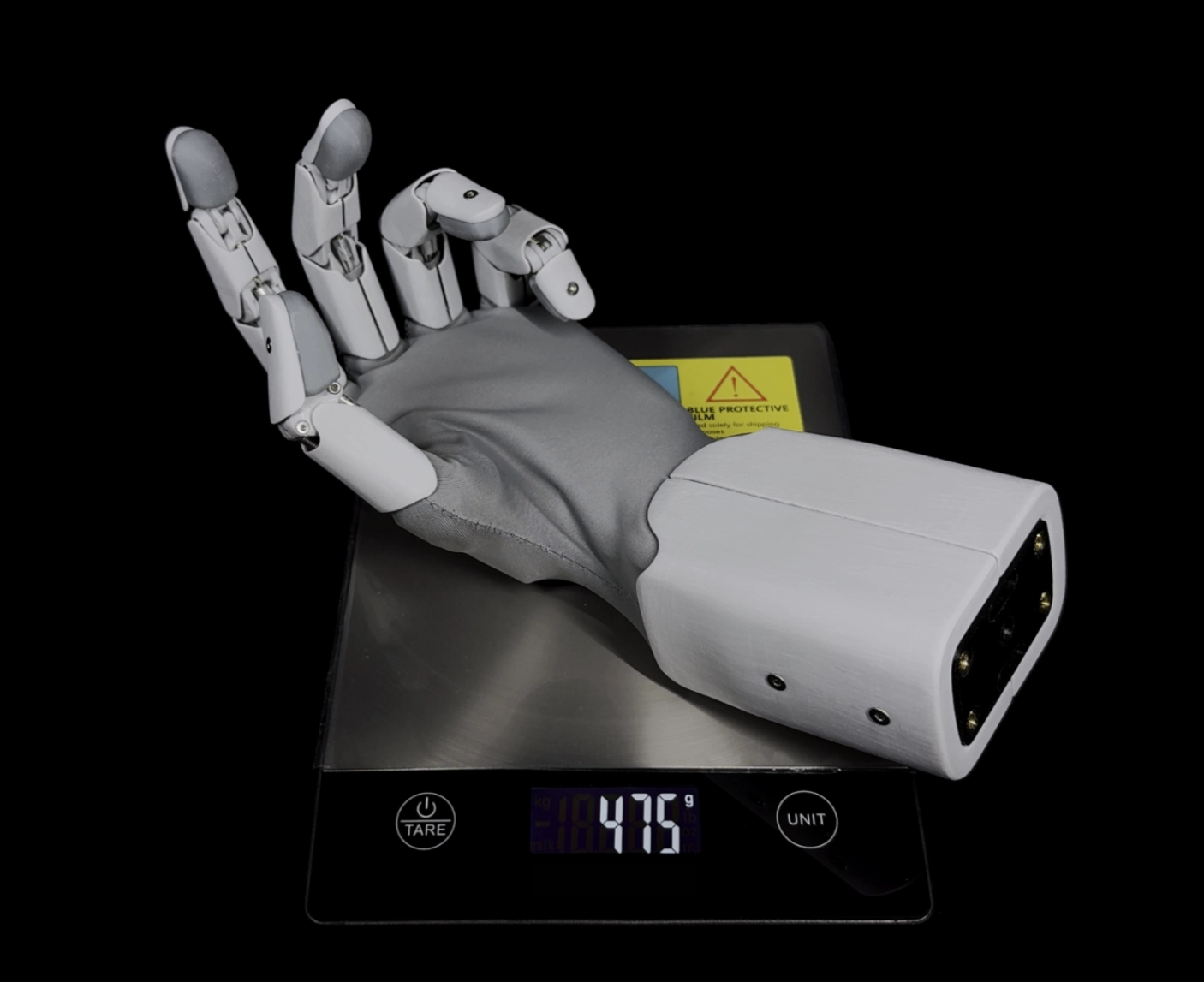 Robotická ruka položená na nerezové digitální váze, která ukazuje hodnotu 475 g.