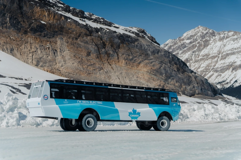První elektrický Ice Explorer na světě, účelově postavený pro ledovcovou turistiku, zachycený z profilu na ledu pod skalnatým masivem na ledovci Athabasca v národním parku Jasper.