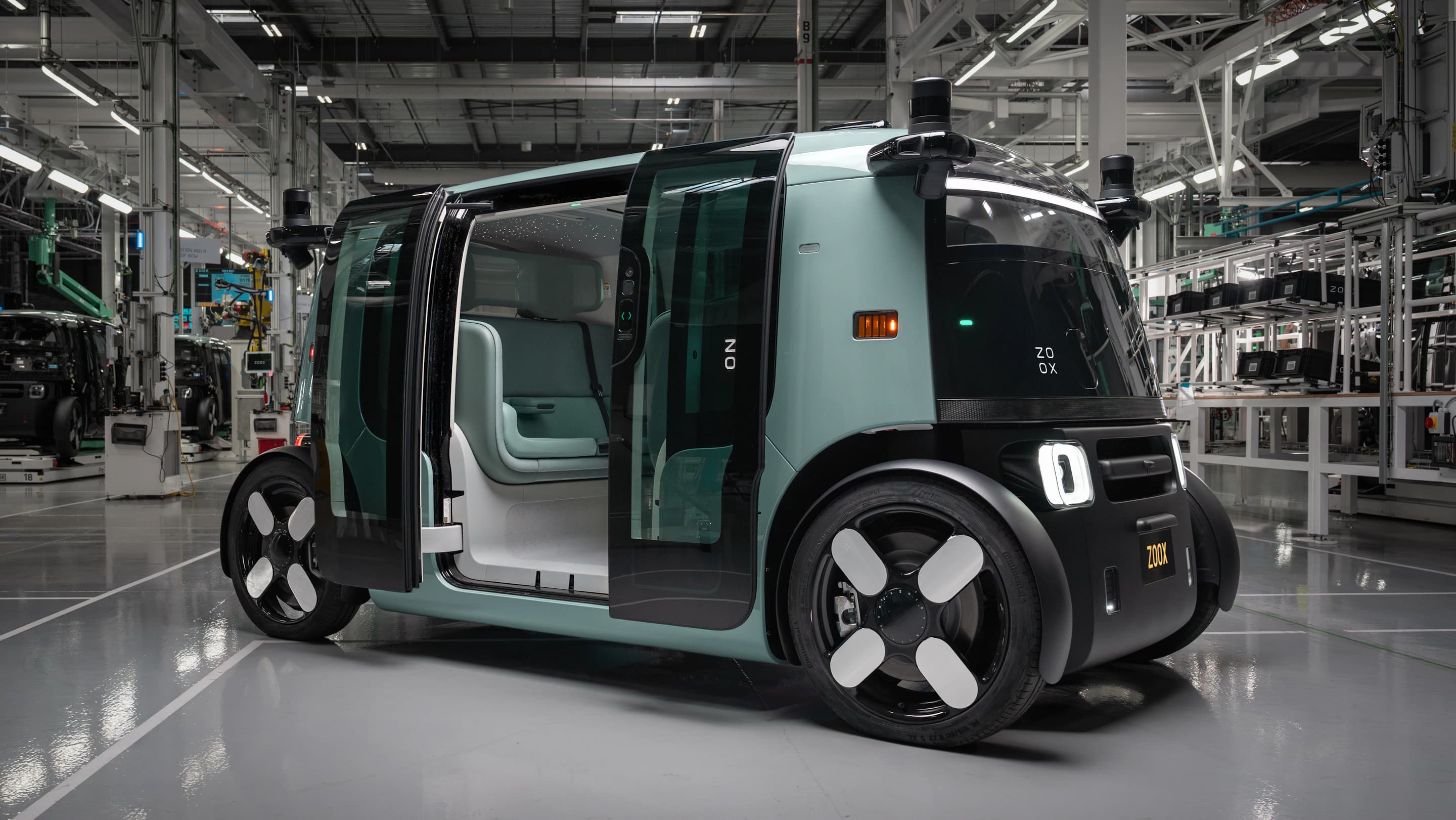 Zelené robotaxi Zoox s otevřenými dveřmi v moderní výrobní hale, odhalující montážní linku a další vozidla v pozadí.