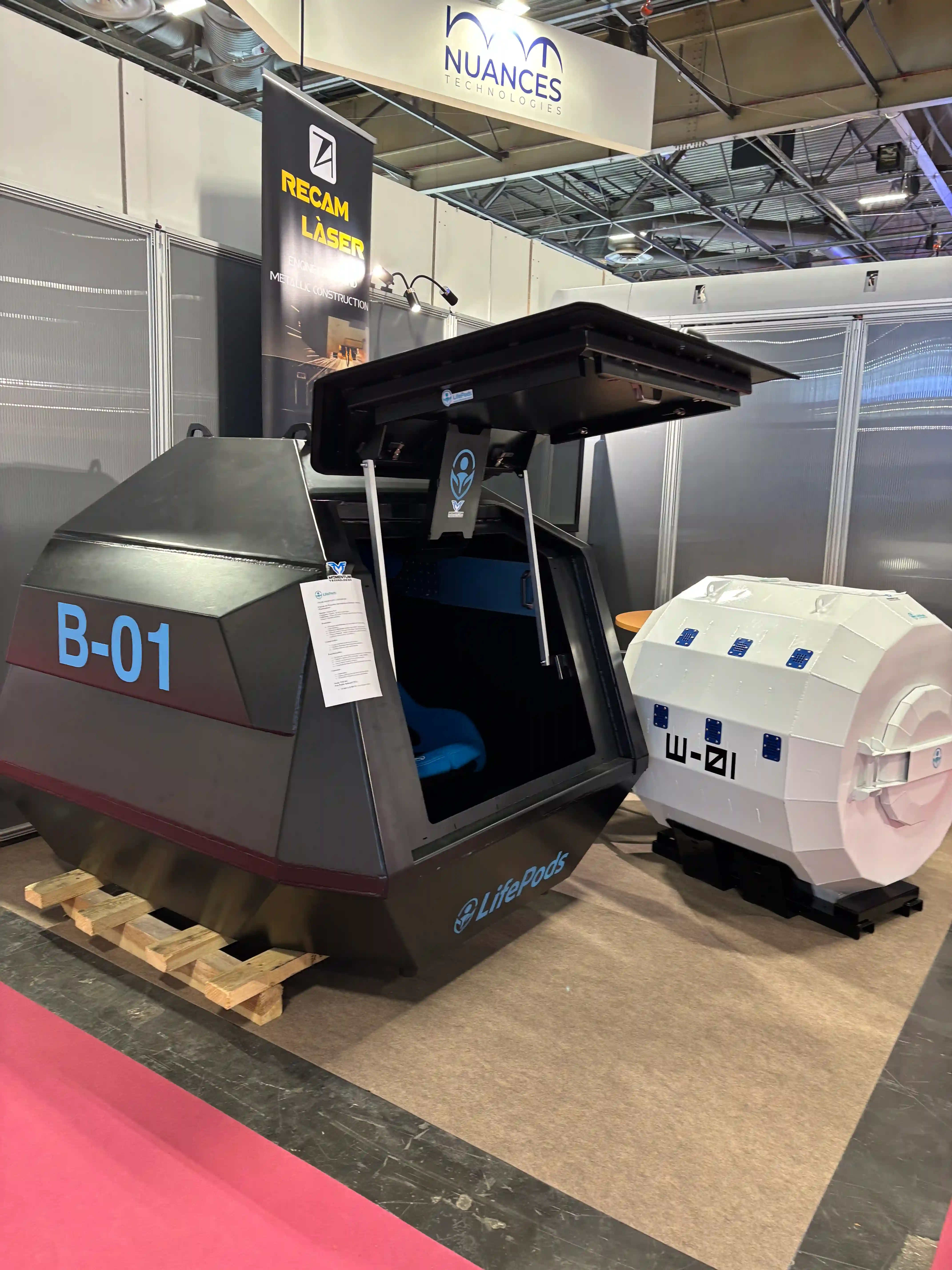 Otevřená černá balistická kapsle LifePods B-01 s modrými sedačkami vedle bílé plovoucí kapsle W-01 na veletržním stánku.
