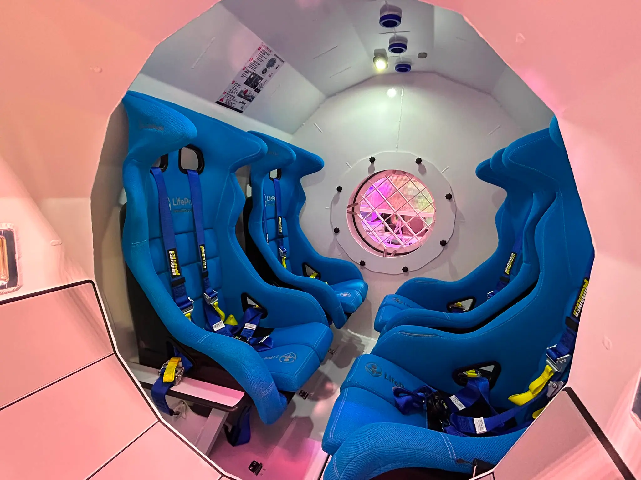 Interiér kapsle LifePods W-01 se čtyřmi modrými sedačkami s bezpečnostními pásy a kulatým oknem s mřížkou.