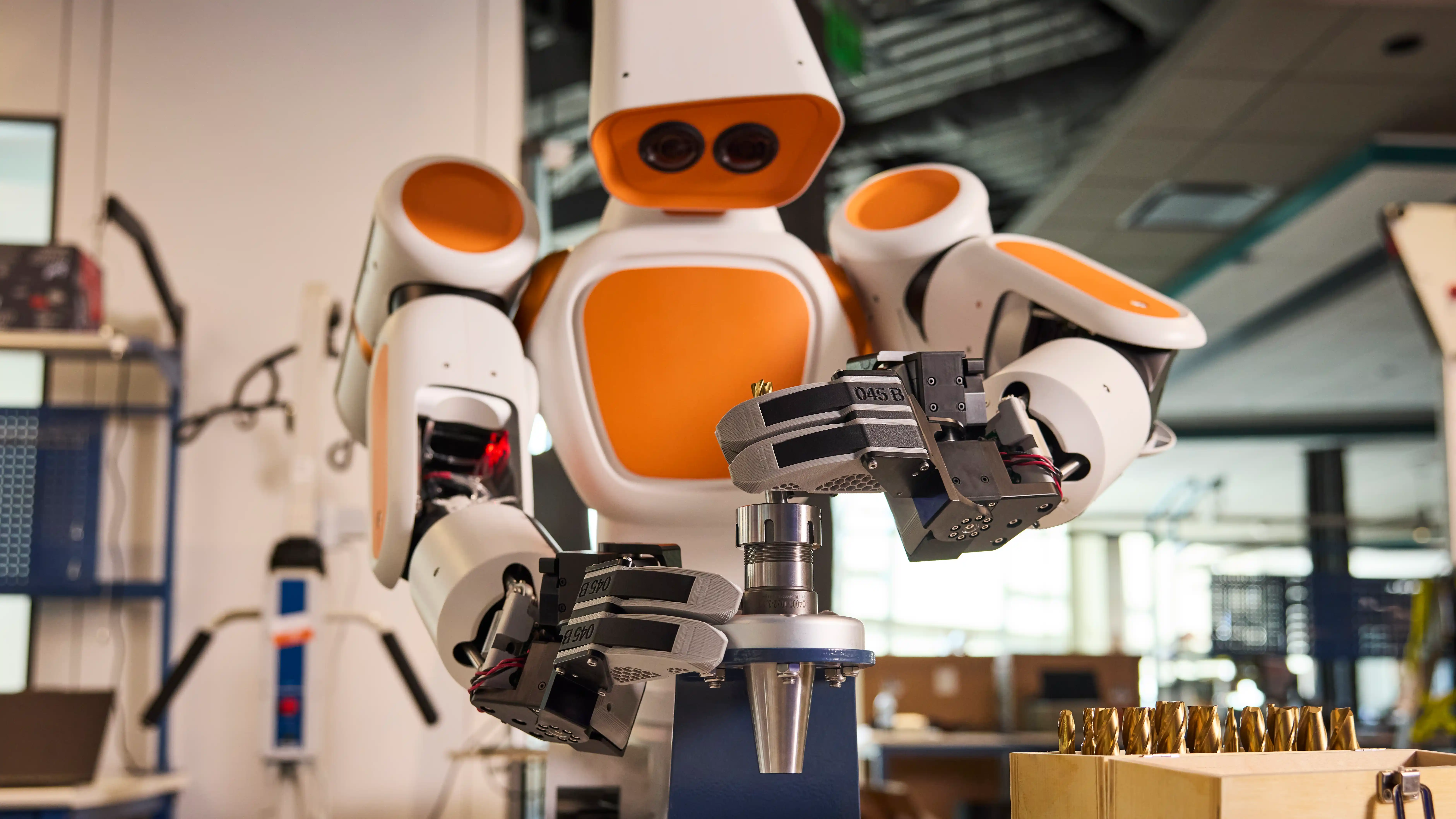 Robot Waldenu drží oběma chapadly kovový nástroj nad upínacím přípravkem v továrním provozu.