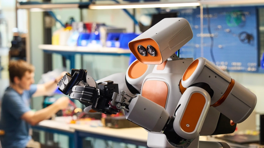 Oranžovobílý robot Waldenu s hlavou se dvěma senzory a chapadlem, v pozadí technik u pracovního stolu.