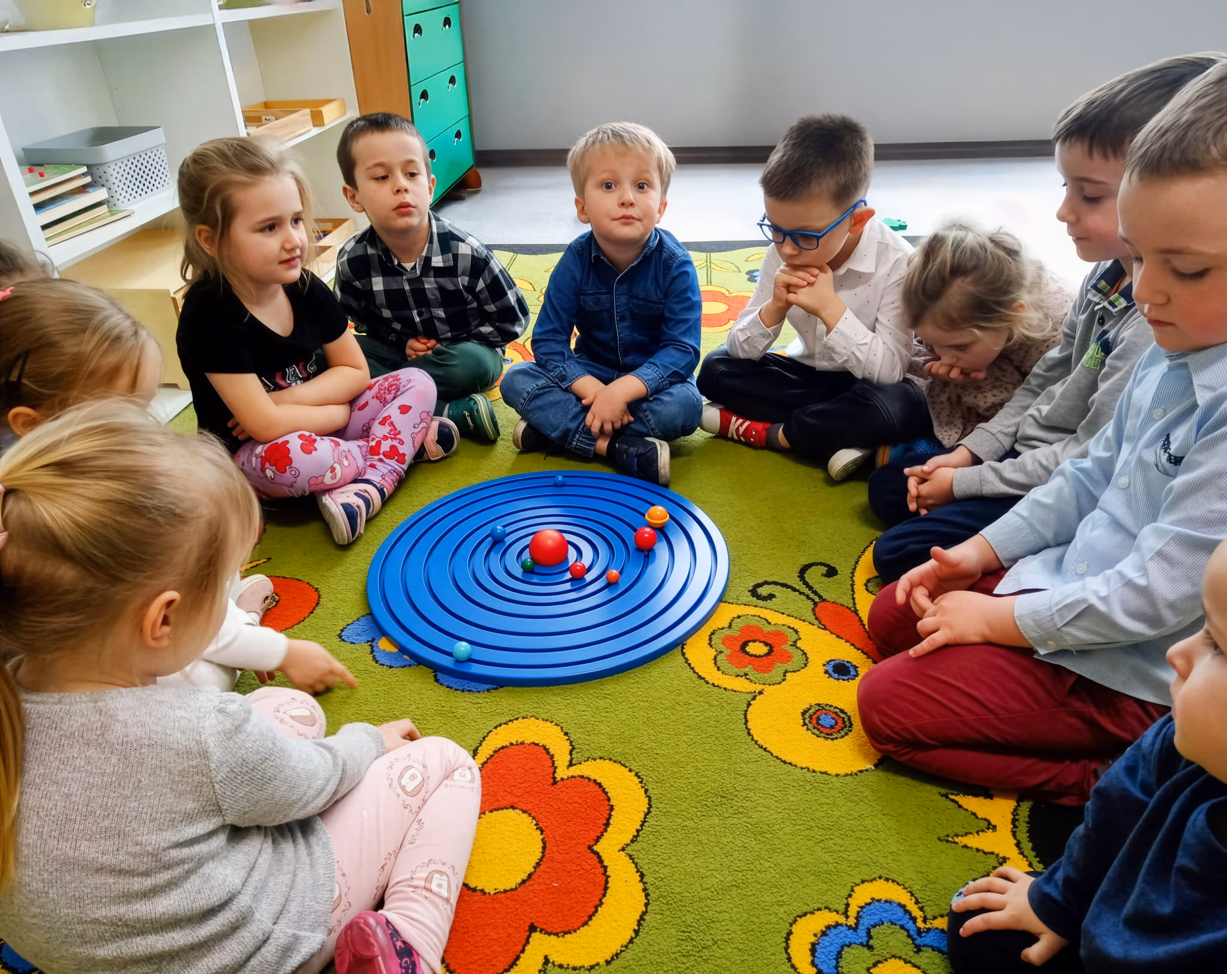Grupa dzieci siedzi w kole na kolorowym dywanie.