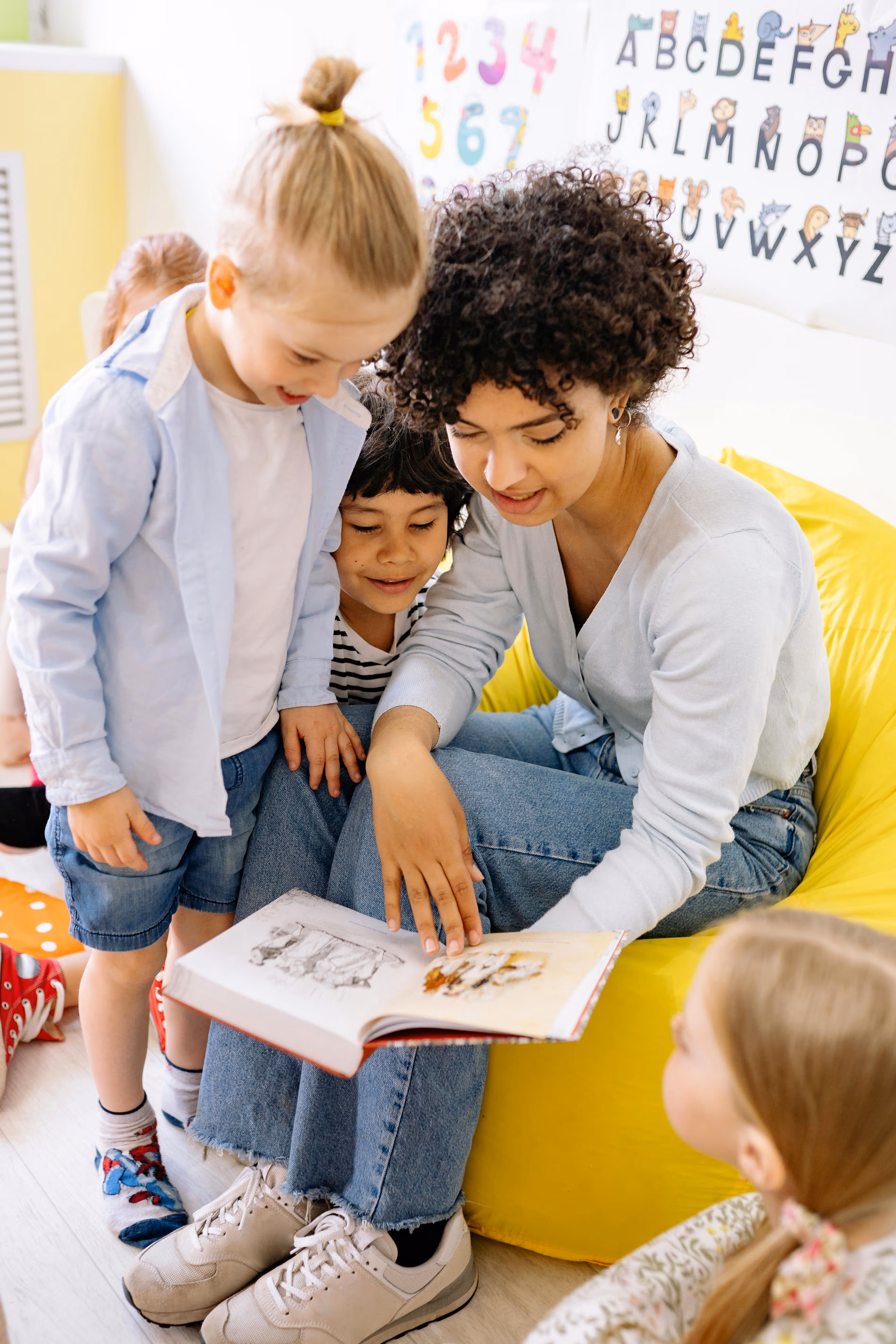 Teacher sitting on a yellow bean bag reading a book to a group of children in a classroom with an alphabet and numbers chart on the wall.