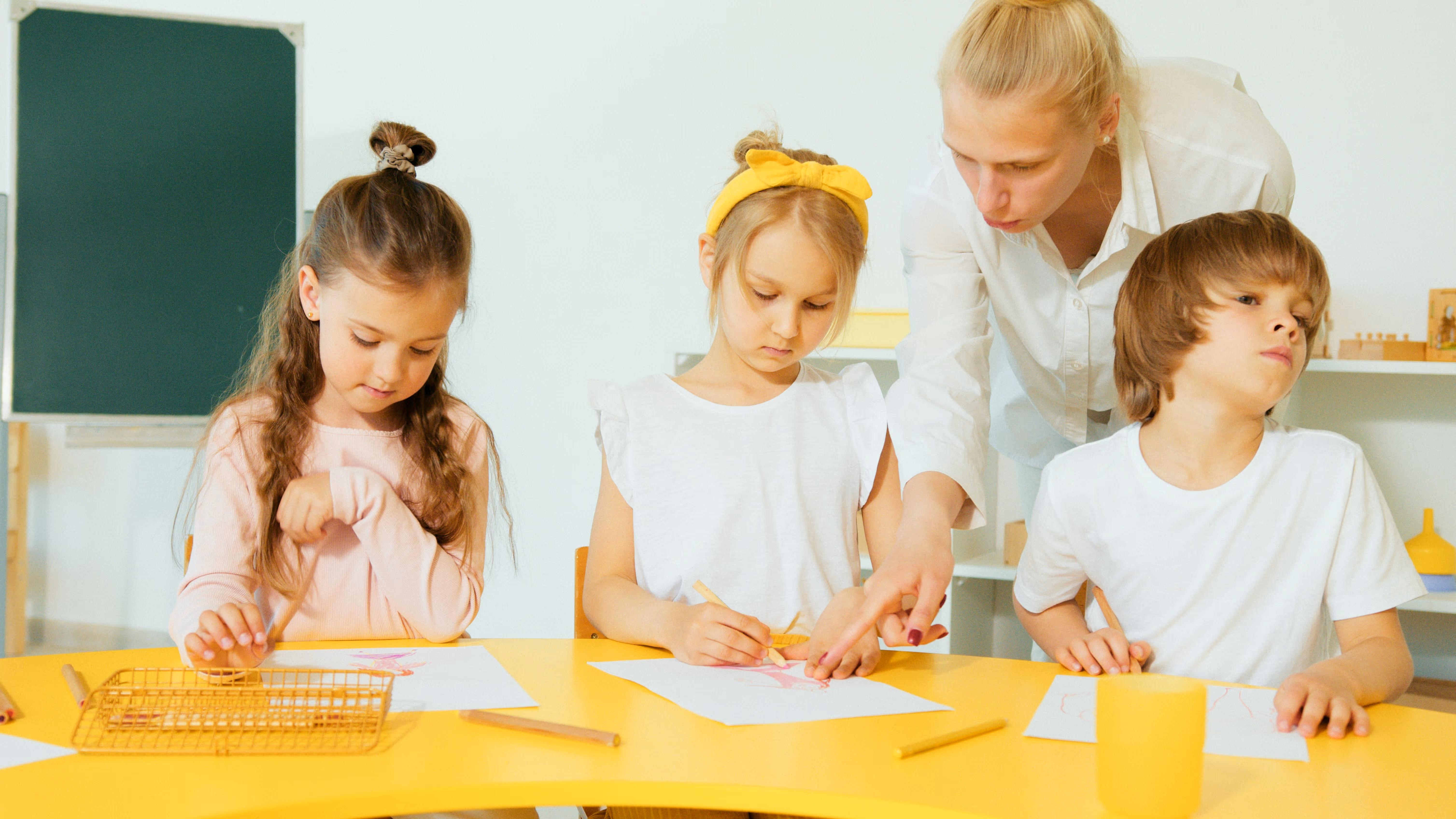 Teacher assisting two girls and one boy drawing with colored pencils at a yellow table in a classroom.