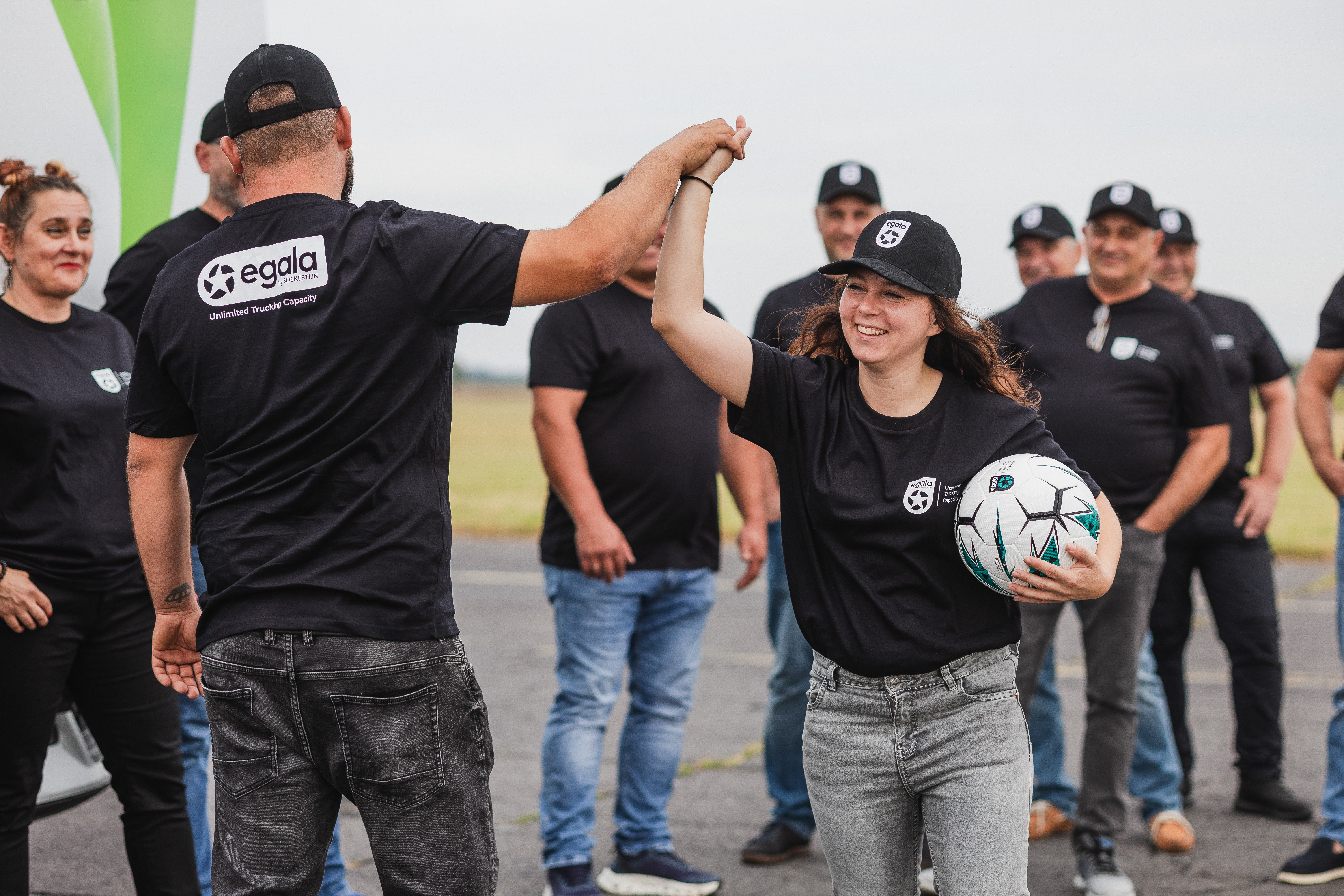 Membră a echipei Egala ținând o minge de fotbal și salutând un coleg, alături de echipă, în cadrul unui eveniment.