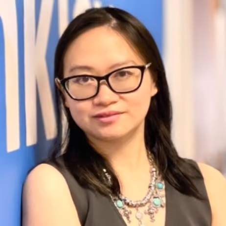 Woman with black hair and glasses wearing a sleeveless top and a turquoise necklace, standing against a blue wall with white text.