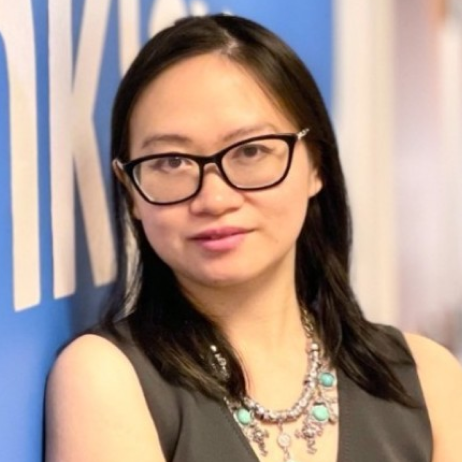 Woman with black hair and glasses wearing a sleeveless top and a turquoise necklace, standing against a blue wall with white text.