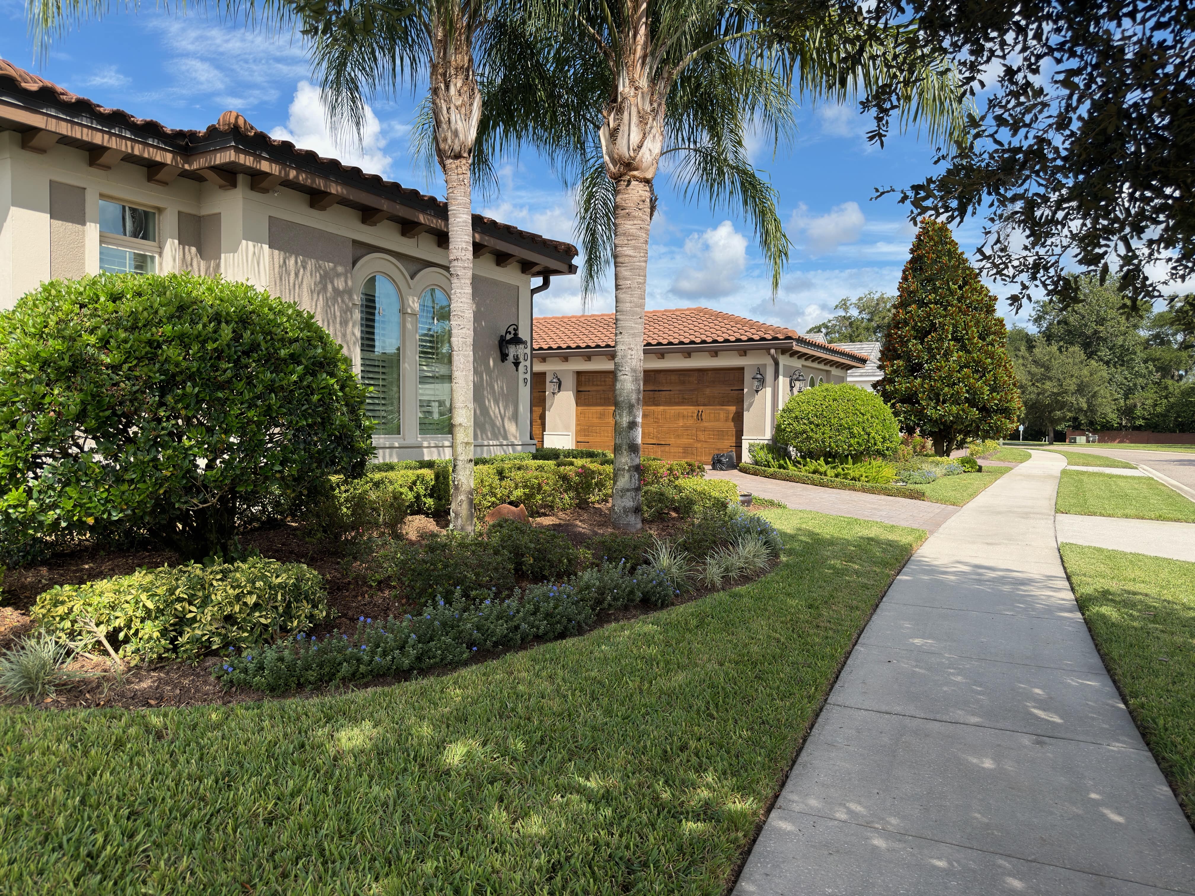 Residential house with beige walls, tiled roof, wooden garage doors, surrounded by palm trees and manicured bushes along a concrete sidewalk.