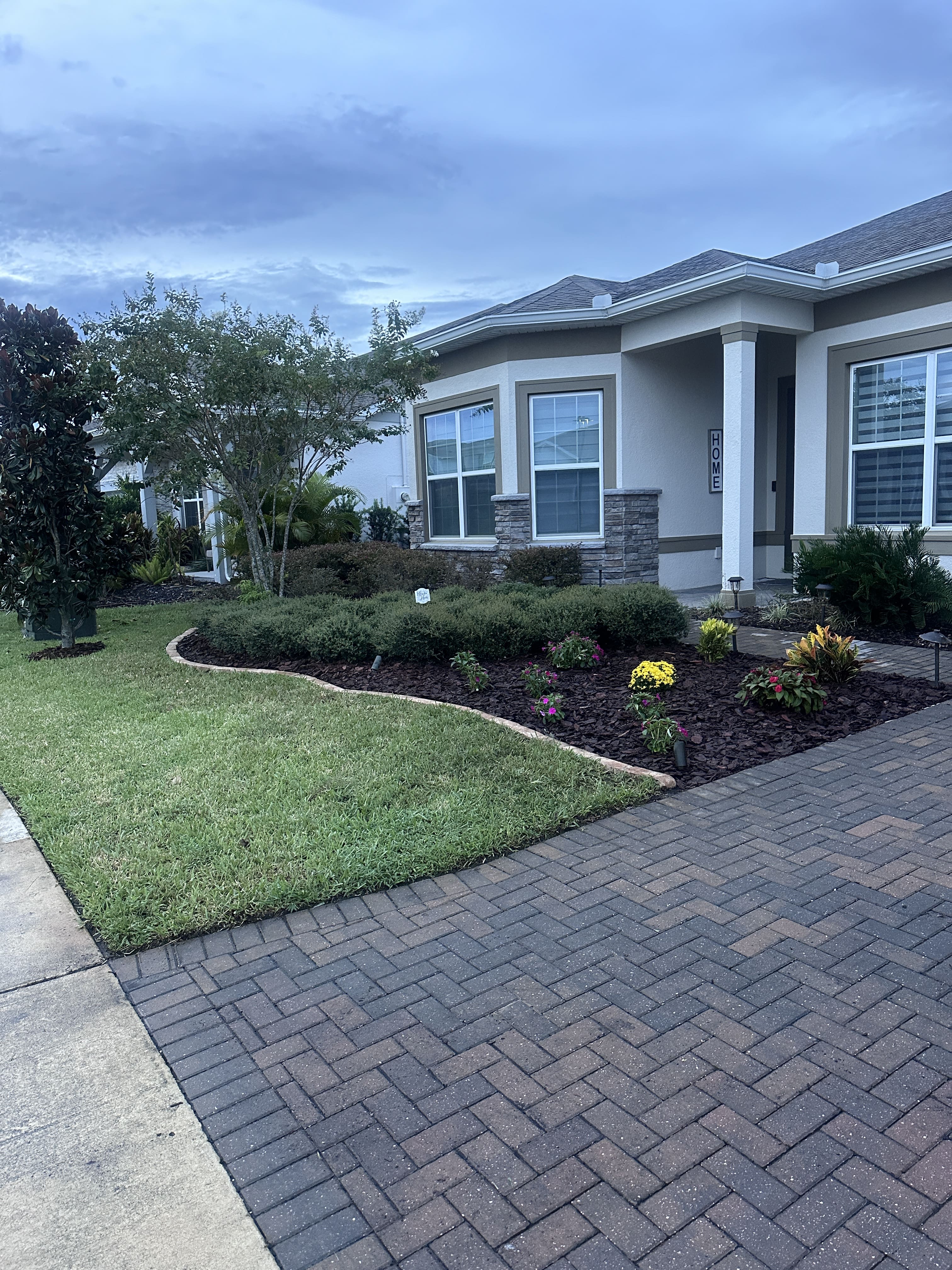 Front yard of a suburban house with a paved driveway, green lawn, and landscaped garden with shrubs and flowers.