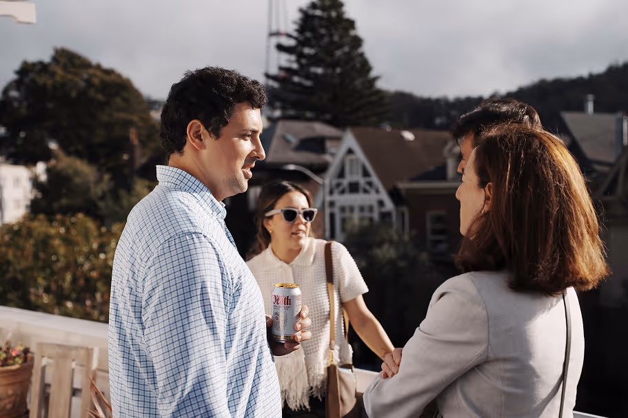 Three people conversing on an outdoor balcony with houses in background