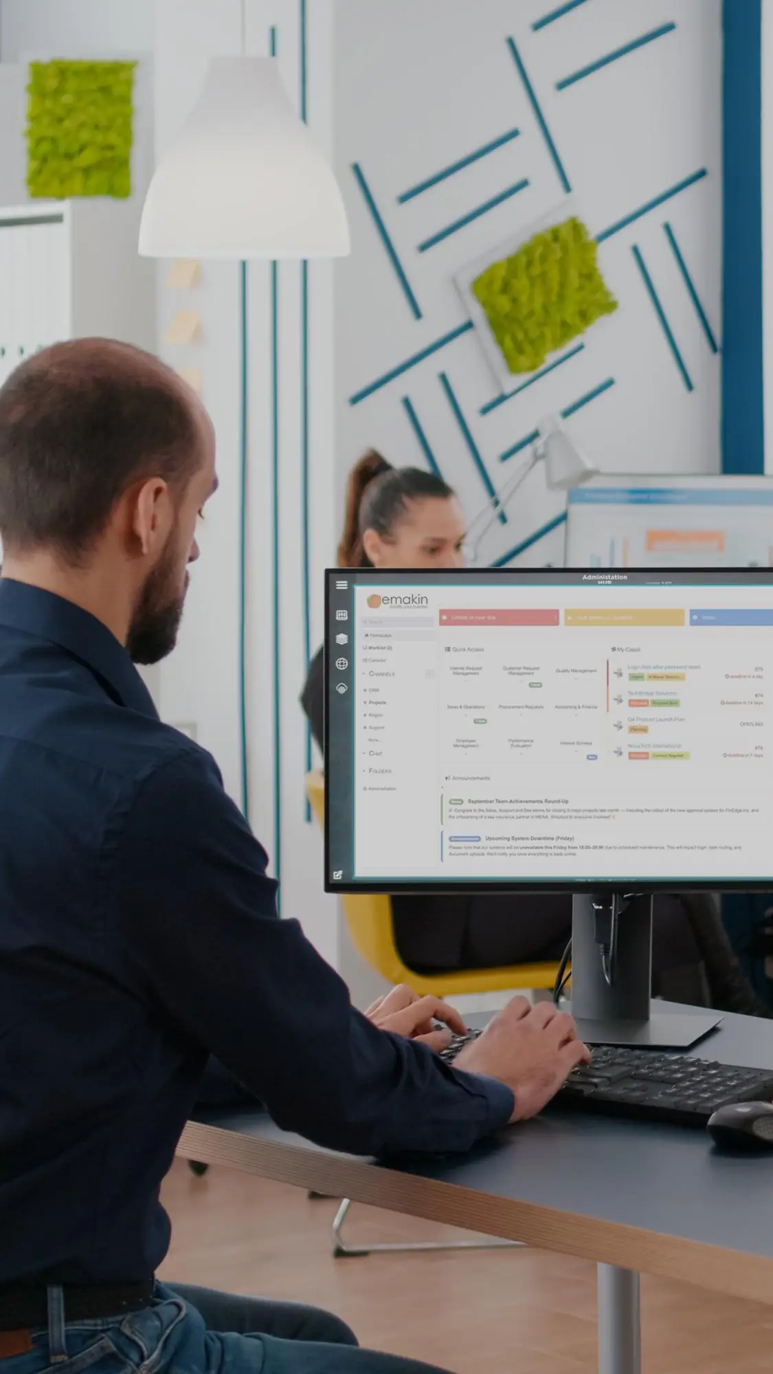 Man in dark blue shirt typing on a keyboard at a desk, with a computer monitor displaying the Emakin software interface in a modern office setting.