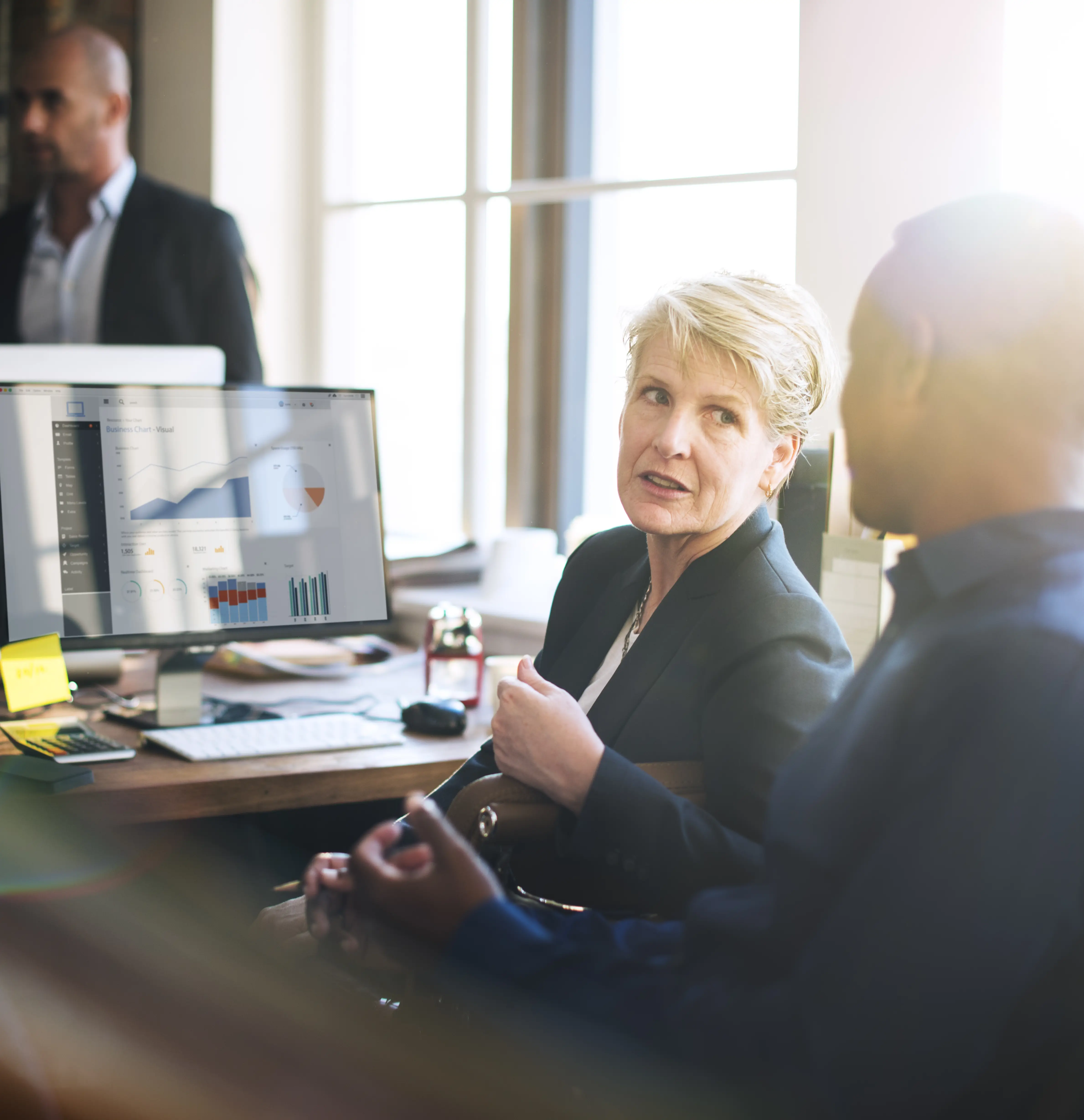 Woman and man in business attire discussing data charts displayed on a computer screen in an office with bright natural light.