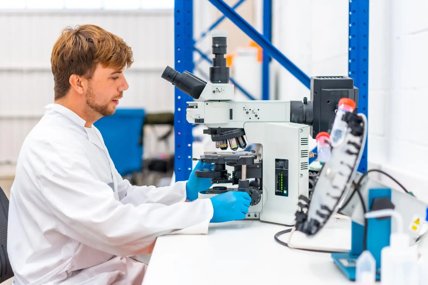 Scientist wearing a white lab coat and blue gloves adjusting a microscope in a laboratory.
