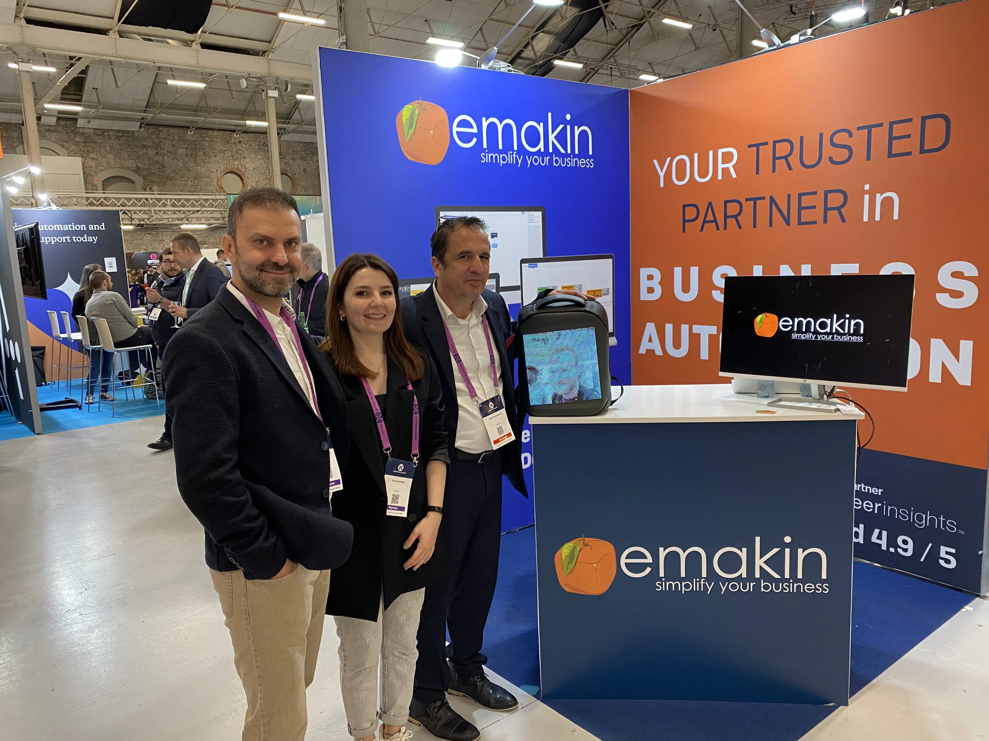 Three people standing and smiling in front of an Emakin business booth with computer monitors and a backpack display.