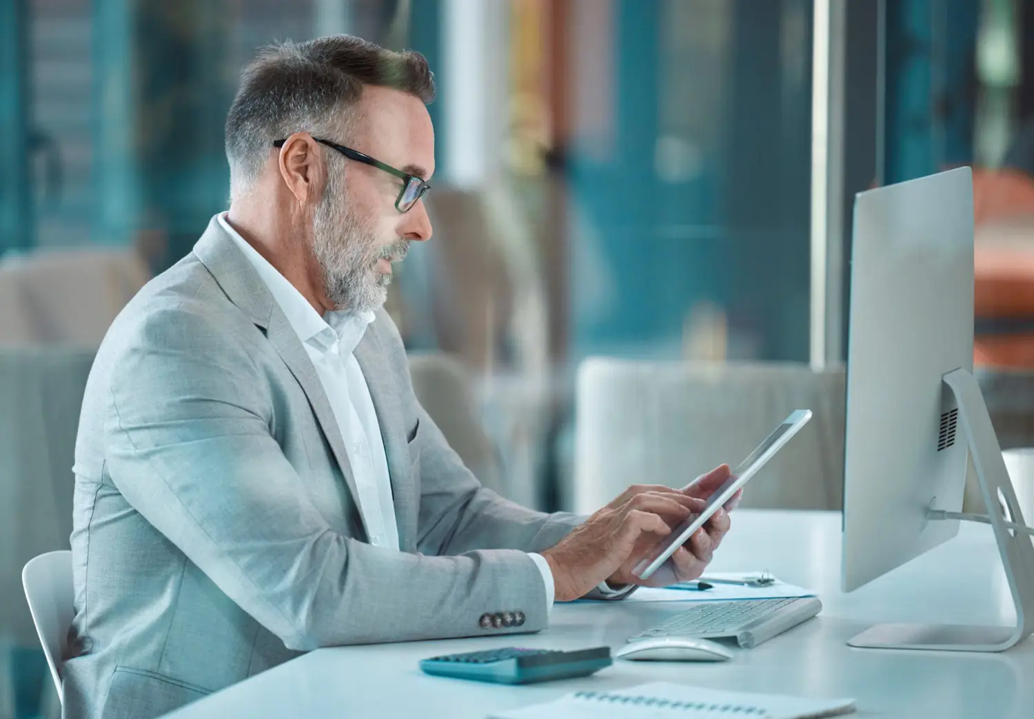 Middle-aged man with glasses in a light gray suit using a tablet at a desk with a computer.