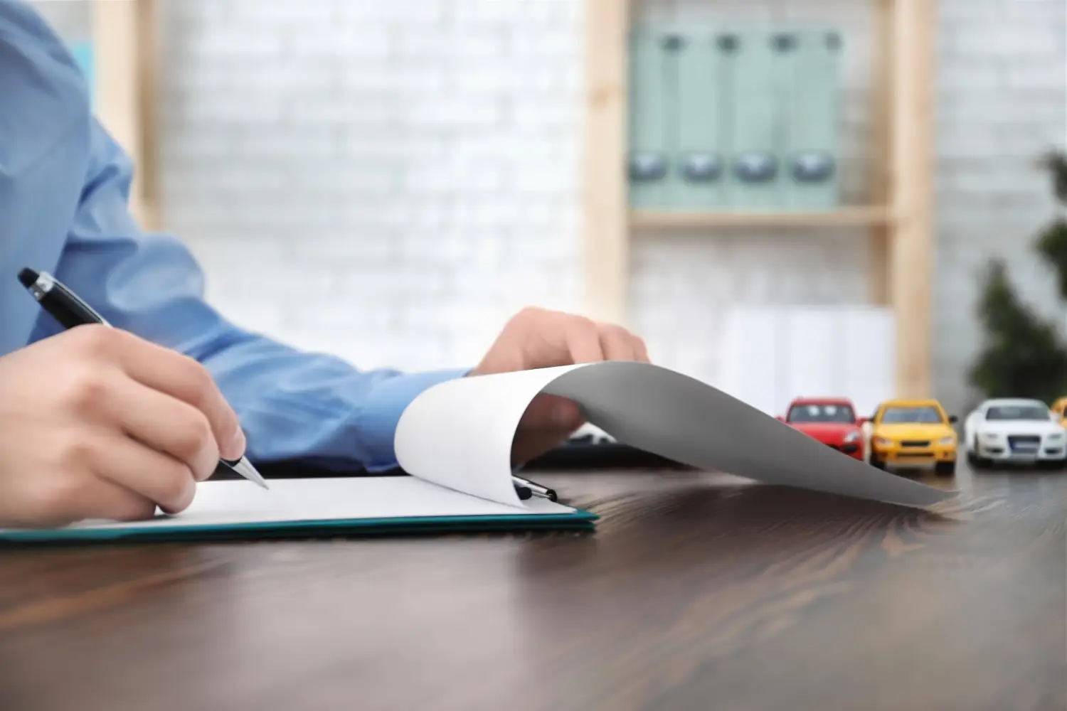 Person in blue shirt writing on paper with a pen, with toy cars lined up on the desk in the background.