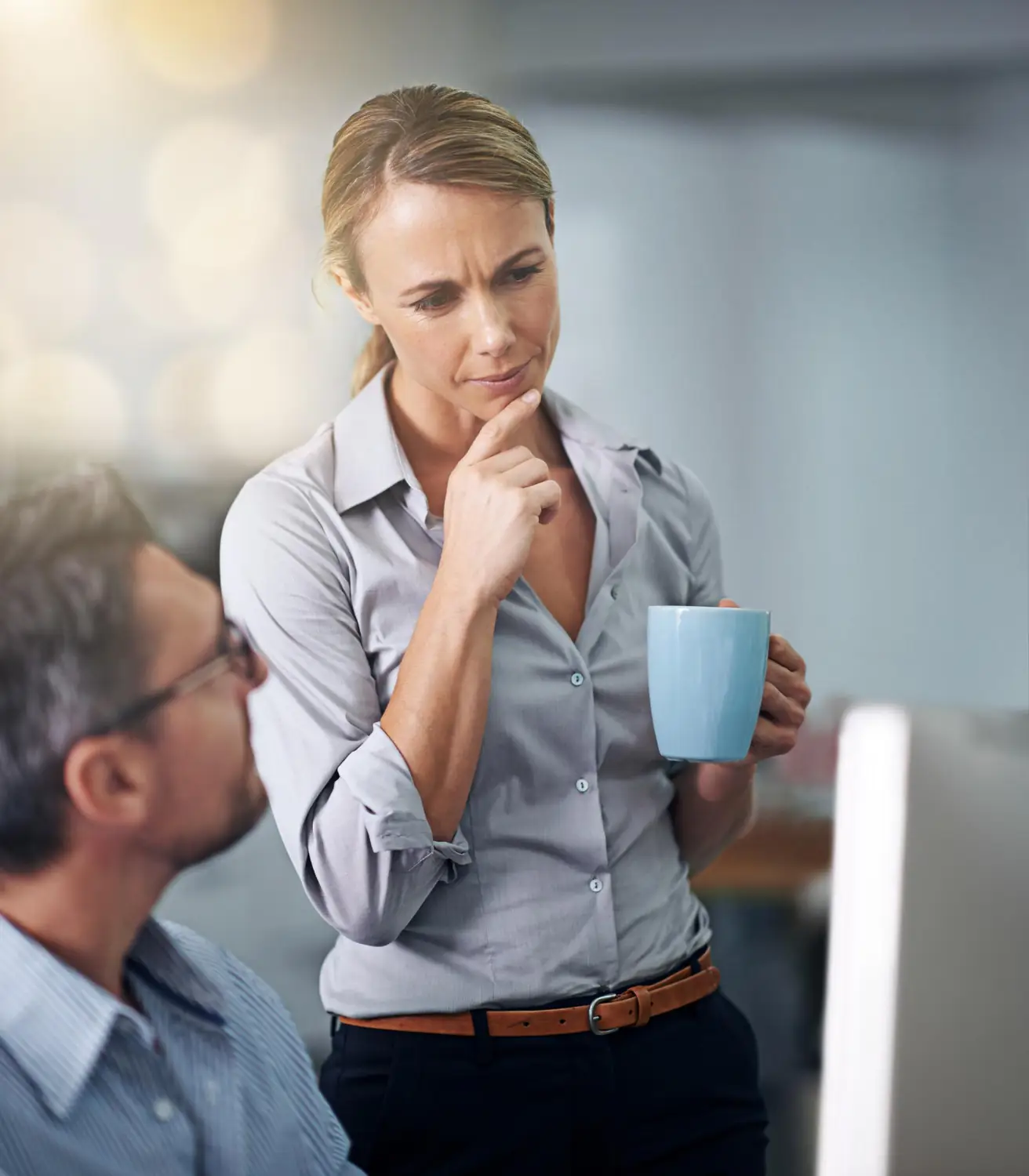 A thoughtful woman holding a blue mug looks at a computer screen while talking to a seated man with glasses in an office setting.