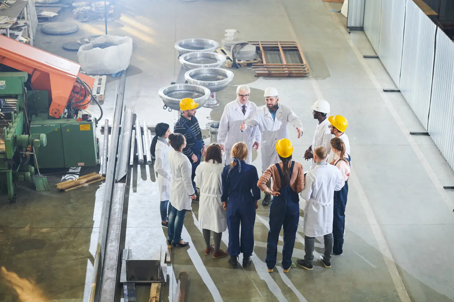 Group of engineers and factory workers in safety helmets and lab coats having a discussion inside an industrial warehouse.