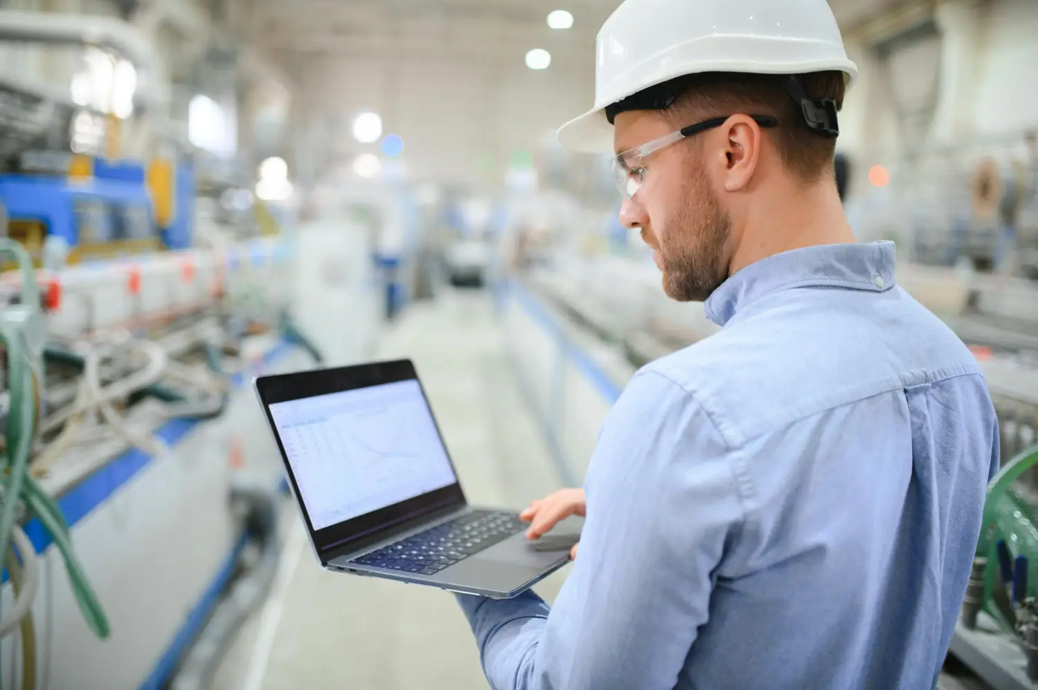 Engineer wearing a white hard hat and safety glasses, working on a laptop in an industrial factory setting.