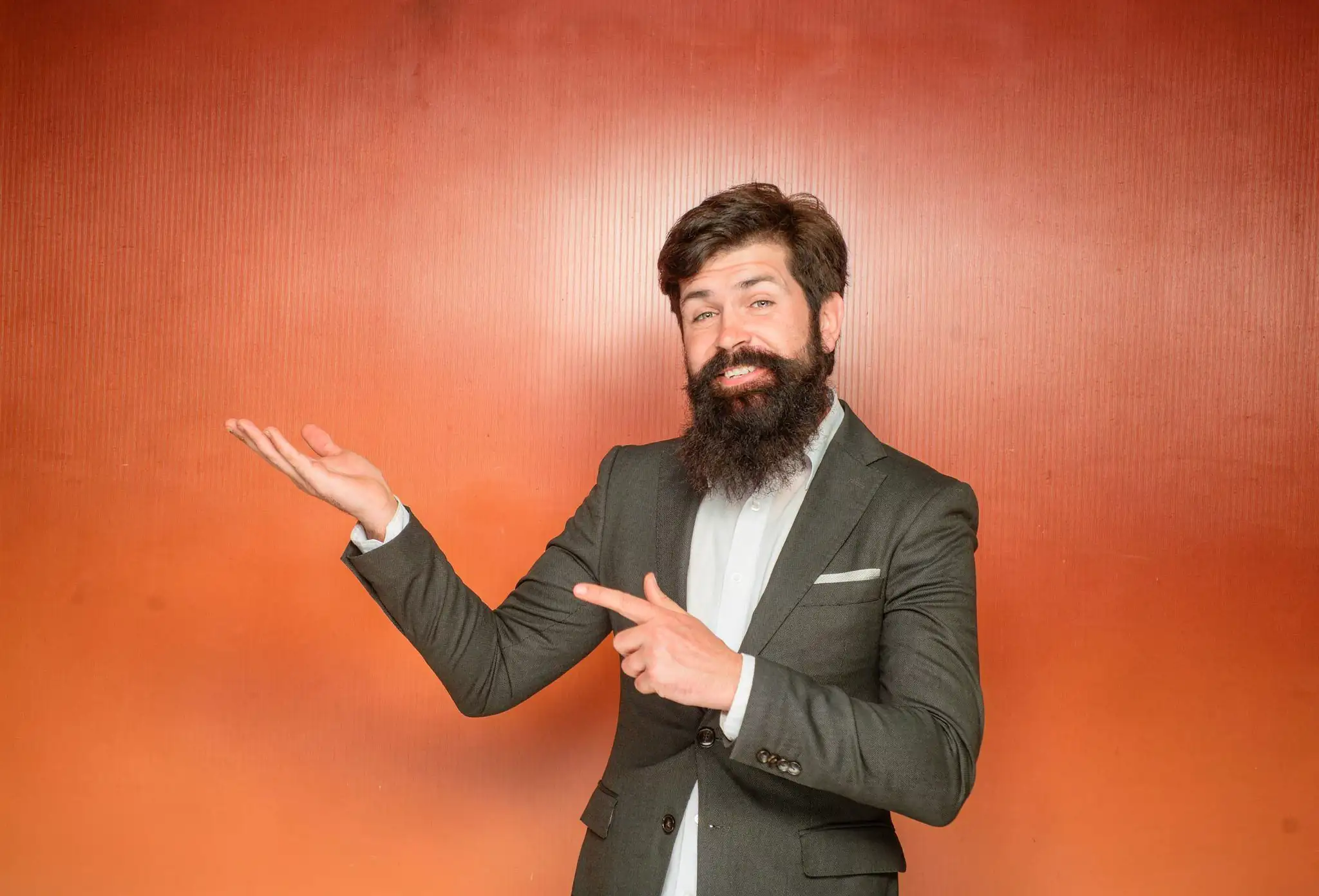 Bearded man in a suit pointing and presenting with one hand against an orange background.