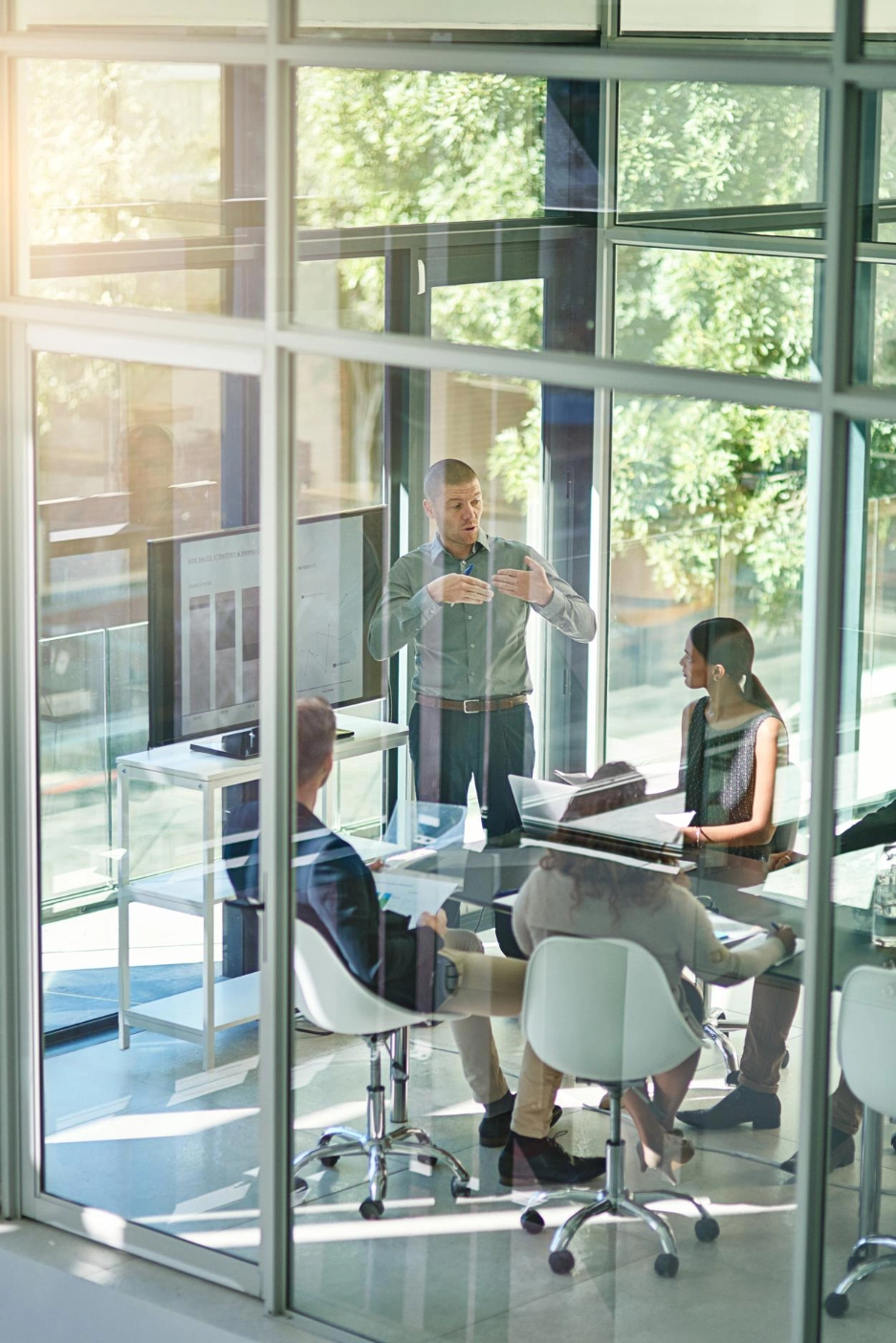 Businessman presenting to colleagues in a glass-walled office with charts on a screen and bright natural light.