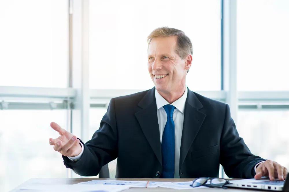 Smiling middle-aged man in a suit and blue tie gesturing while sitting at a desk with papers, glasses, and a laptop in a bright office.