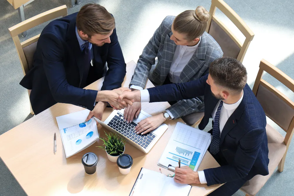 Three business professionals sitting around a table with a laptop and documents, two men shaking hands over a successful meeting.