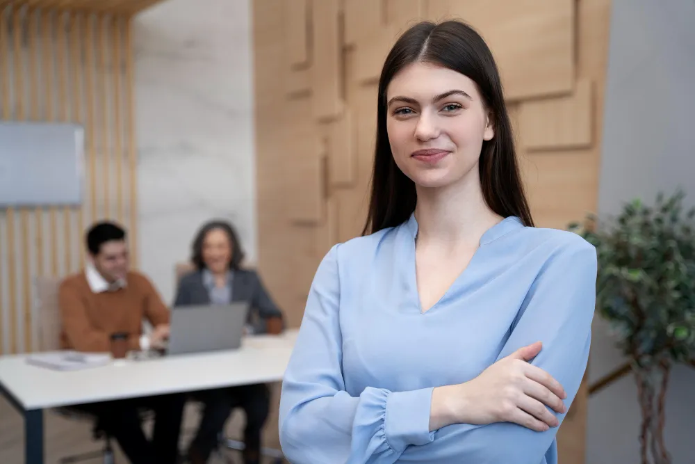 Confident young woman in a light blue blouse standing with arms crossed in a modern office, with two colleagues working at a table in the background.