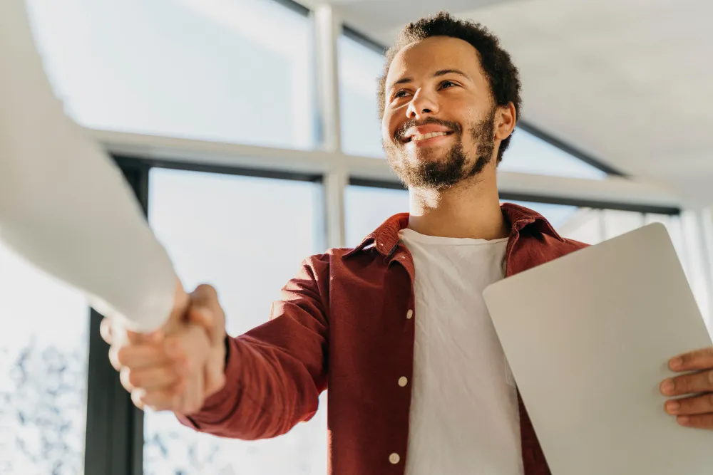 Smiling man holding a laptop shaking hands with another person in a bright office.