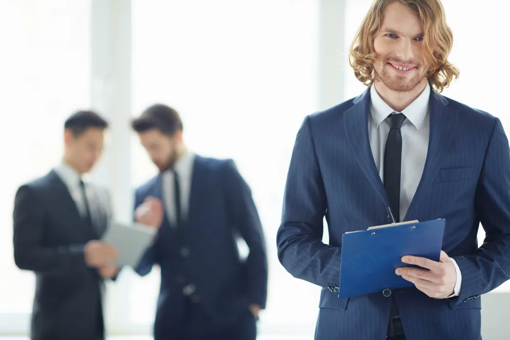 Smiling young businessman in a suit holding a clipboard with two colleagues blurred in the background.