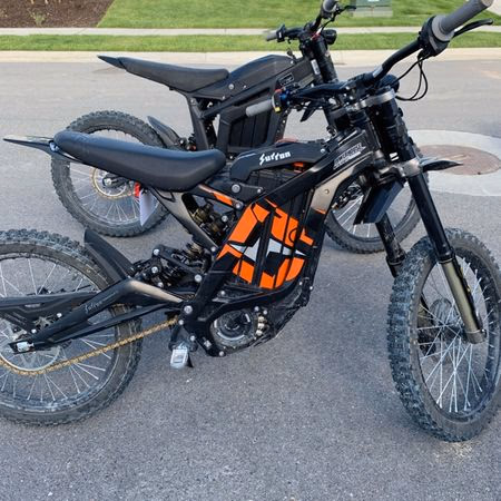 Two black and orange electric dirt bikes parked on a paved surface outdoors.