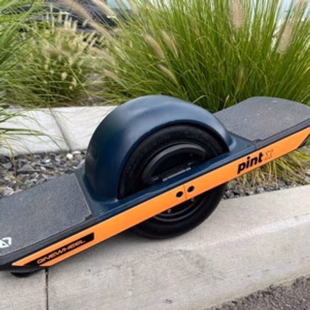 One-wheeled electric skateboard with black and orange design resting on a concrete curb near green plants.