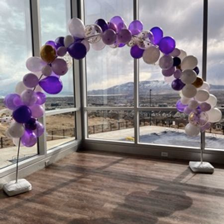 Indoor purple and white balloon arch with gold accents in front of large windows overlooking a mountainous landscape.