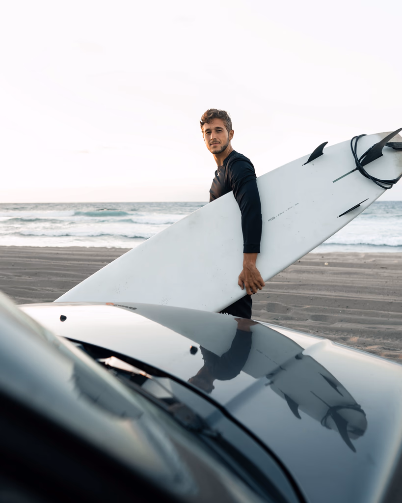 Man in black wetsuit holding a white surfboard near a car by the beach with ocean waves in the background.