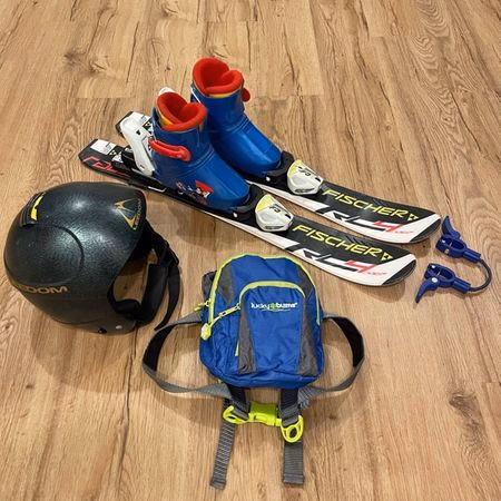Pair of Fischer RC4 skis with attached blue and red ski boots, black ski helmet, and blue kids' backpack on a wooden floor.