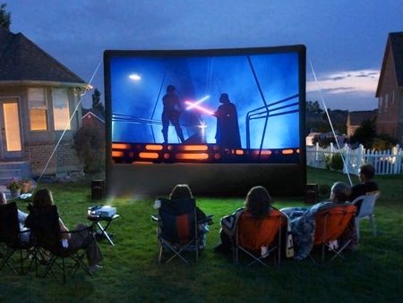 Group of people seated on lawn chairs watching an outdoor movie screen showing a lightsaber duel scene at dusk.