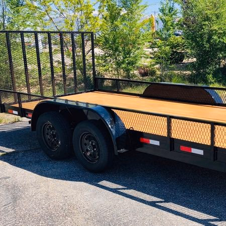 Flatbed utility trailer with two black wheels and metal mesh sides parked on a paved road with trees in the background.