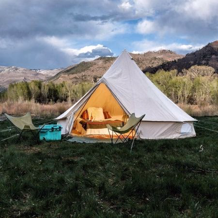 Lit white bell tent with cozy interior and pillows set up on green grass near mountains under a cloudy sky.
