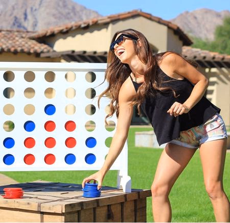 Smiling woman in sunglasses placing a blue disc on a large outdoor Connect Four game in a sunny backyard.