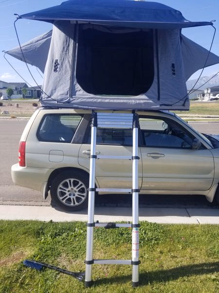 Silver SUV parked on a street with a grey rooftop tent and a ladder extending to the ground.