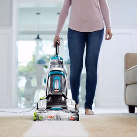 Person vacuuming a carpet with a turquoise upright vacuum cleaner in a bright living room.