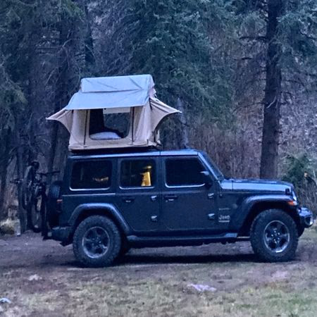 Dark green Jeep Wrangler with rooftop tent set up in a forested camping area.
