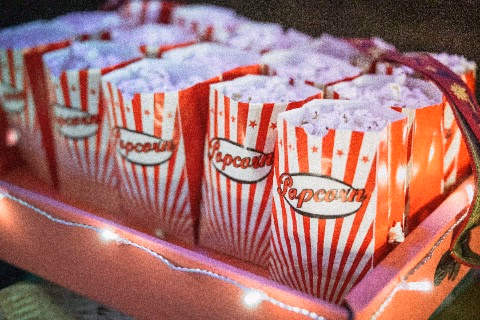 Several red and white striped popcorn bags filled with popcorn arranged in a box with string lights around it.