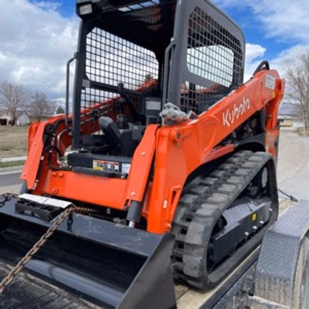 Red Kubota compact track loader on a trailer under a partly cloudy sky.