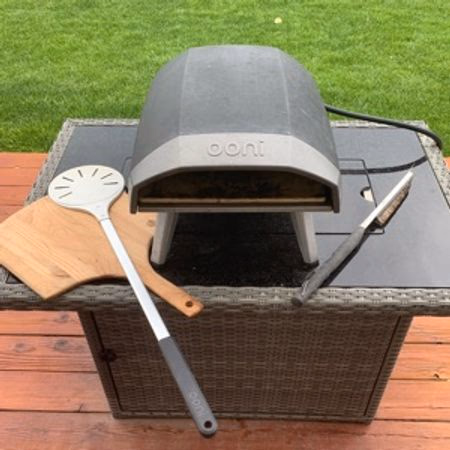 Outdoor Ooni pizza oven on a wicker patio table with a wooden pizza peel and a perforated pizza spatula beside it.