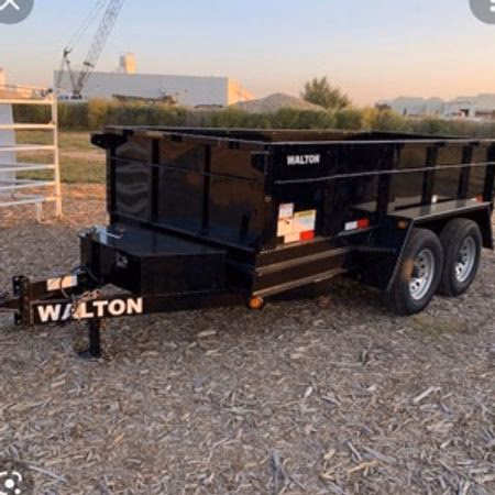 Black Walton dump trailer with dual axles parked on a dirt and wood chip ground near industrial buildings.