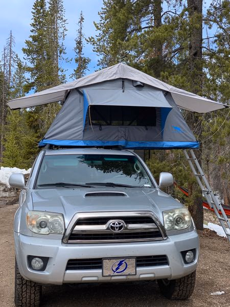 Silver Toyota SUV parked outdoors with a blue and gray rooftop tent set up on its roof, surrounded by tall evergreen trees.